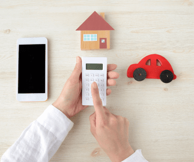 A hand holds a calculator surrounded by a small car, house and phone