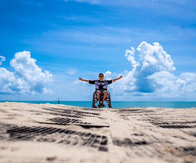 A man in a wheelchair with his arms outstretched on a beach with a blue sky in the background