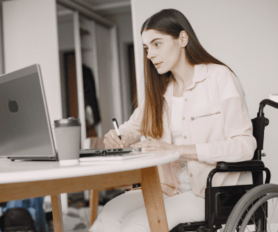 A woman in a wheelchair sits at a desk and looks at a laptop