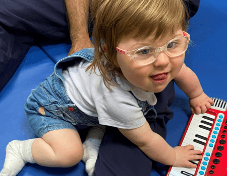 A little girl with fair hair and glasses, sitting on the floor, smiling, and playing a red toy keyboard